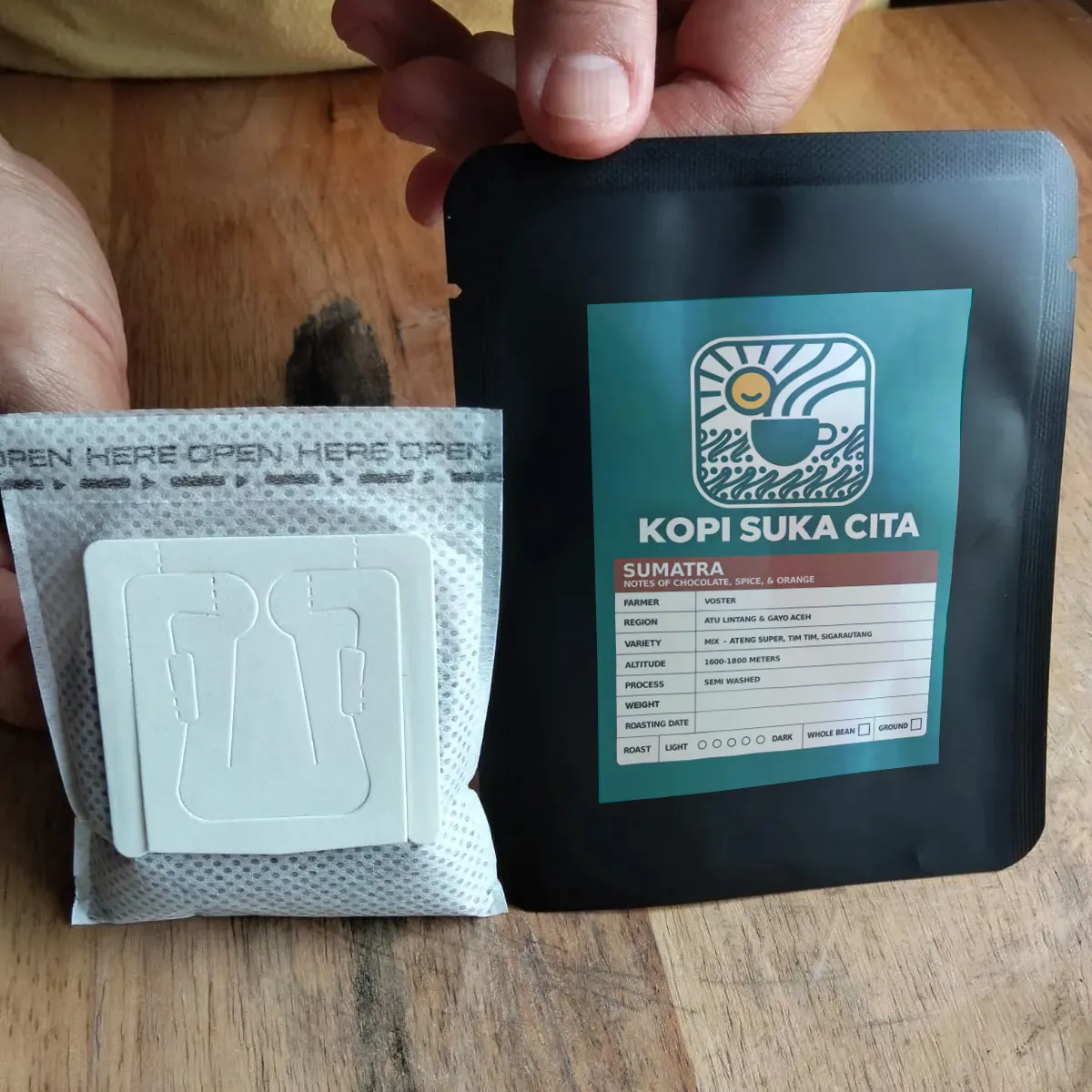 A Kopi Suka Cita single-brew drip bag next to the whole-bean pack it comes from